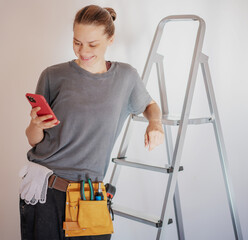 Beautiful young happy woman repairman standing by a stepladder in a belt with tools on a white wall...