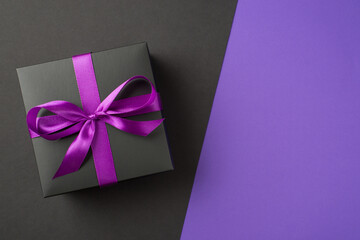 Above photo of black giftbox with purple ribbon wrapped as bow isolated on the half violet and half black divided abstract background