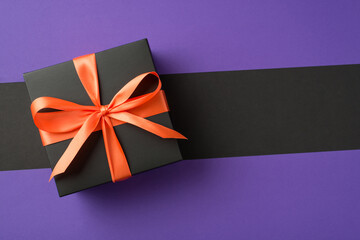 Top view photo of black giftbox with orange ribbon bow on isolated violet and black background with copyspace