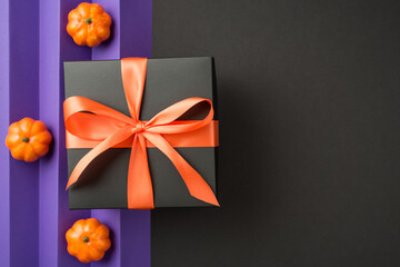 Top view photo of halloween composition black giftbox with orange ribbon bow and small pumpkins on isolated back and violet sheet with vertical folds background with copyspace