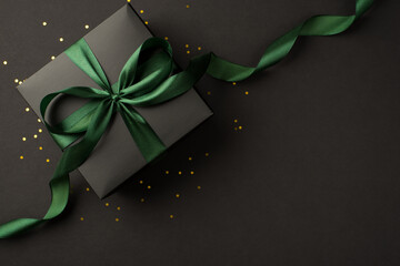 Top view photo of stylish black giftbox with green satin ribbon bow and golden confetti on isolated black background with empty space