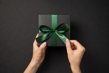 First person top view photo of female hands unpacking black giftbox with green satin ribbon bow on...