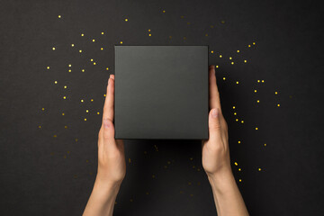 First person top view photo of woman hands holding black giftbox and golden sequins on isolated...