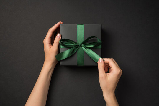 First person top view photo of hands unpacking black giftbox with green satin ribbon bow on isolated black background