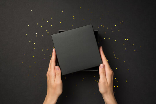 First person top view photo of hands holding open lid on black giftbox and golden sequins on isolated black background with empty space