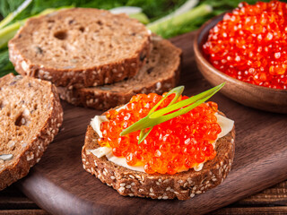 Sandwich with red caviar on a wooden tray surrounded by bread, caviar and herbs, horizontal