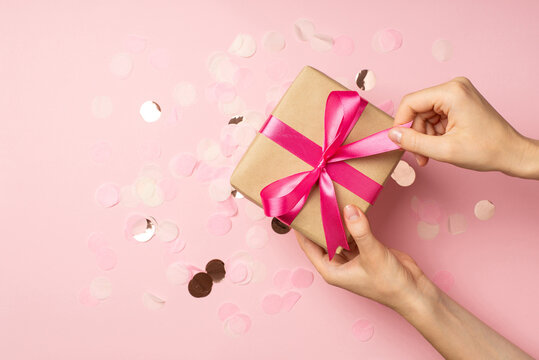 First person top view photo of hands unpacking craft paper giftbox with pink ribbon bow over large shiny sequins on isolated pastel pink background with copyspace