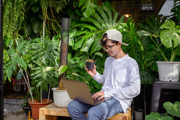 Handsome Asian man loves to plant trees sitting in a garden monstera is using his laptop computer to market and sell trees Handsome Asian man is interested in doing business selling trees online.