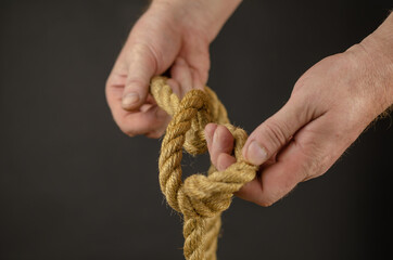 Man ties knot against black background. Hands holding yellow jute rope. Close-up. Selective Focus.