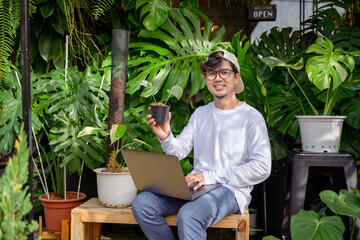 Naklejka na ściany i meble Handsome Asian man loves to plant trees sitting in a garden monstera is using his laptop computer to market and sell trees Handsome Asian man is interested in doing business selling trees online.
