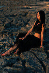 Young slim sexy girl with long straight dark hair, dressed in long black dress sits on the stones barefoot and looks forward, at the sea in Sicily, south Italy
