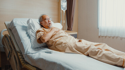 Sad senior Asia man having having heart attack lying on hospital bed and press emergency button. Sick aged guy lying hospitalized in a medical clinic. Health insurance or hospitality concept.