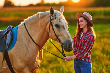 Young cute happy joyful satisfied smiling cowgirl with beautiful blond palomino horse at meadow at...