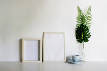 Two frames with coffee and monstera leave on a white wall with nature light. 