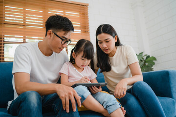 Happy Asian family dad, mom and daughter using computer tablet technology sitting sofa in living room at house. Self-isolation, stay at home, social distancing, quarantine for coronavirus prevention.