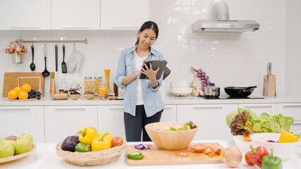 Happy Asian woman using tablet for looking recipe while making food in the kitchen, female use organic vegetable for healthy food at home. Lifestyle women making food concept.