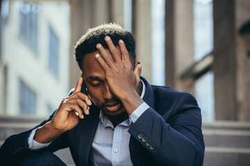 Depressed african american businessman telling sad news talking on cell phone sitting on office...