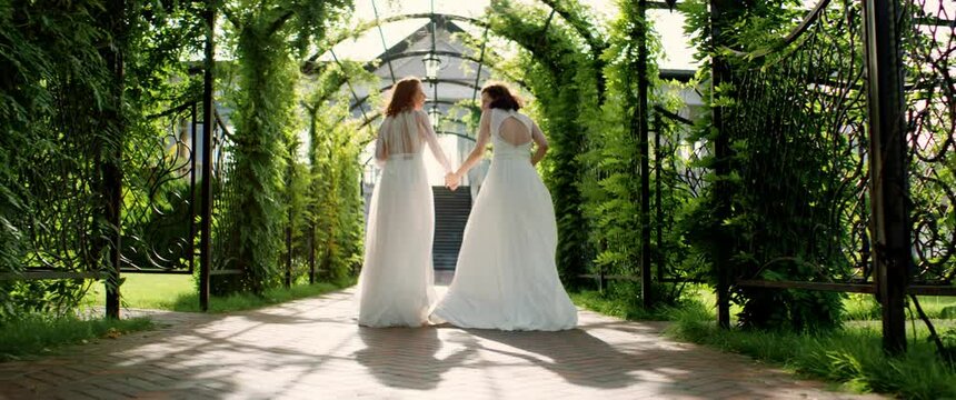 TRACKING Candid shot of two happy lesbian LGBT brides wearing boho dresses walking through alley on their wedding day. Shot with 2x anamorphic lens