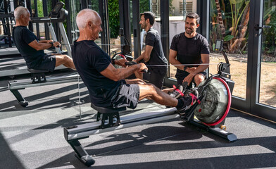 An elderly European man, in a gym, with a personal trainer, exercising simulating rowing, on a simulator, indoor.