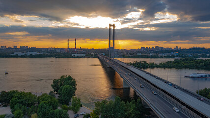 South bridge in Kiev. Sunset over the Dnieper. Thick clouds over the evening city. Evening shot of the bridge. Orange sun at sunset. The rays break through the clouds and are reflected in the river.
