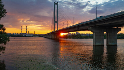 Fototapeta na wymiar South bridge in Kiev. Sunset over the Dnieper. Thick clouds over the evening city. Evening shot of the bridge. Orange sun at sunset. The rays break through the clouds and are reflected in the river.