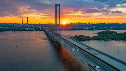 Fototapeta na wymiar South bridge in Kiev. Sunset over the Dnieper. Thick clouds over the evening city. Evening shot of the bridge. Orange sun at sunset. The rays break through the clouds and are reflected in the river.