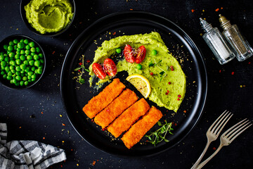 Fried fish sticks with potato and peas puree on wooden black table
