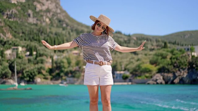 Young happy carefree woman in straw hat standing on turquoise sea bay background and enjoying vacation. Girl imagine that she is bird. Travel destination, amazing nature, south Europe.