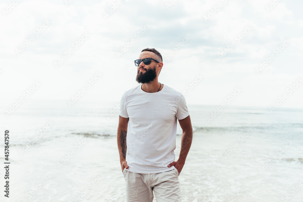 Sticker Young bearded hipster guy with beard in white t-shirt and sunglasses at the tropical beach. Mock-up for print. T-shirt template.