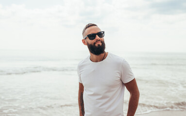 Young bearded hipster guy with beard in white t-shirt and sunglasses at the tropical beach. Mock-up for print. T-shirt template.
