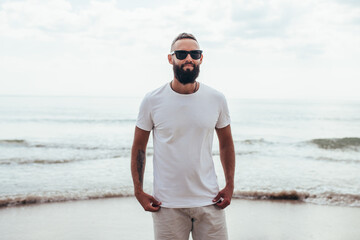Young bearded hipster guy with beard in white t-shirt and sunglasses at the tropical beach. Mock-up...