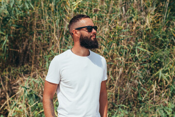 Naklejka na ściany i meble Young bearded hipster guy with beard in white t-shirt and sunglasses at the tropical beach. Mock-up for print. T-shirt template.