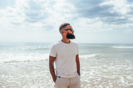 Young bearded hipster guy with beard in white t-shirt and sunglasses at the tropical beach. Mock-up for print. T-shirt template.