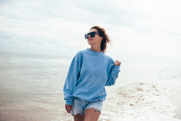 Beautiful young adult woman in blue blank sweatshirt and sunglasses on a background of a tropical...