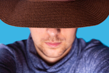 Close up photo of a man making selfie in the brown hat putting his head down and hiding his face over the blue background.