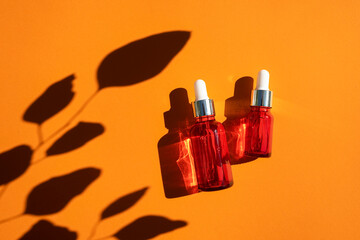 Red glass dropper bottle with silver lid on an orange background. Skincare products with beautiful daylight. Trending concept natural cosmetic with eucalyptus leaves shadow. Beauty concept for face