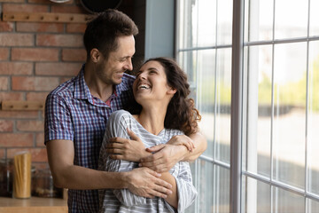 Affectionate happy young man cuddling smiling attractive wife, standing near huge window in own...