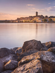 view through stones and lake to city Marta on lake Bolsena in Italy in morning lights - obrazy, fototapety, plakaty