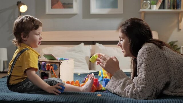 Mom and baby are playing. Positive mother and her little child are sitting on bed in bedroom and playing building blocks. Slow motion