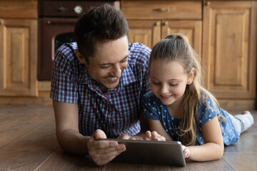 Caring young father using digital computer tablet with little adorable kid daughter, playing games online, watching funny cartoons, lying together on warm heated floor, family tech addiction concept.