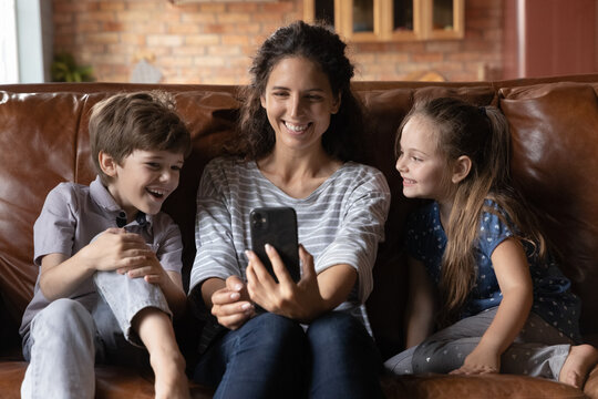Happy beautiful millennial latin mother holding cellphone in hands, recording funny video with adorable little kids siblings, enjoying pleasant web camera communication together or making photos.