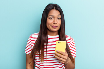 Young Venezuelan woman holding mobile phone isolated on blue background confused, feels doubtful and unsure.