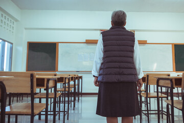 Senior Asian Teacher standing behind alone looking empty classroom no children student when back...