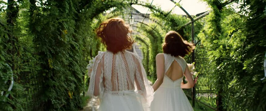 TRACKING Candid shot of two happy lesbian LGBT brides newlyweds running and kissing in the green alley on their wedding day. Shot with 2x anamorphic lens