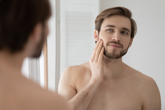 Young shirtless man looking in mirror touch his face after shave applied aftershave cream enjoy smooth, flawless, soft skin. Grooming, morning routine, ad of skincare cosmetic products for men concept