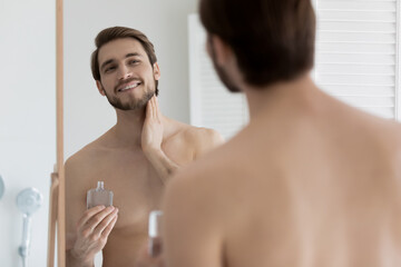 Young 30s handsome shirtless man looking in mirror applying to skin aftershave lotion, standing in...