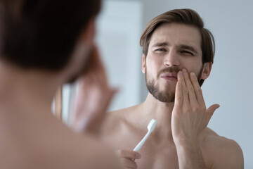 Handsome young man look in mirror touch cheek with hand, feels pain while brushing teeth due cavity...