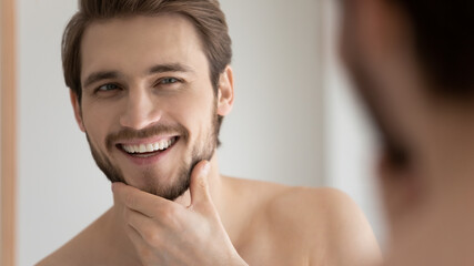 Fototapeta na wymiar Close up view handsome male face reflecting in mirror, touch bristled chin in front of mirror feels satisfied after facial shaving, cream good results. Morning routine, self-hygiene, skincare concept