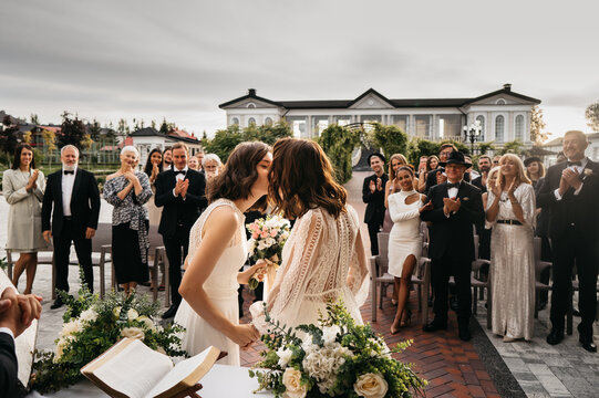 Portrait of two LGBT females lesbians brides kissing during wedding ceremony, guests clapping and cheering. Shot with 2x anamorphic lens