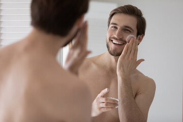 Handsome smiling shirtless man holds cream jar apply moisturiser facial creme, reflects in mirror looks at camera. Lifting nourishing anti-wrinkles treatment for soft hydrated skin. Skincare concept